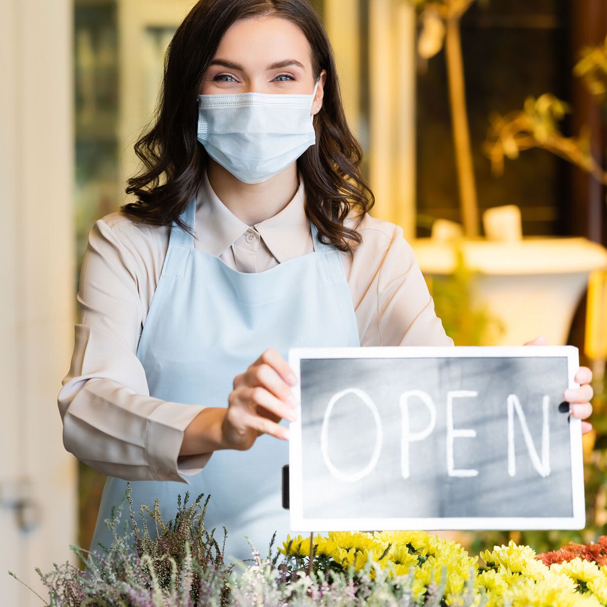 Masked merchant lady holding an OPEN shop sign.